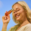Model applying  Vitamin C Glow Potion serum bottle from The Jojoba Company skincare  to their face with a clear blue sky background